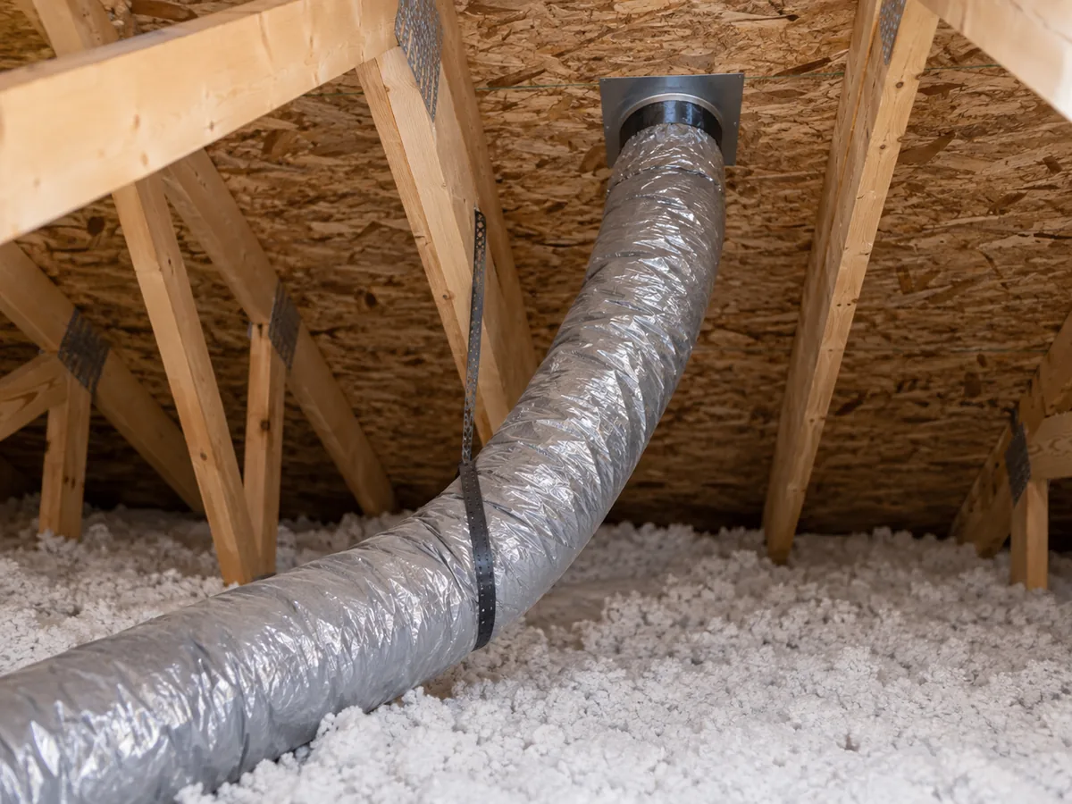 A foil-jacketed exhaust duct routed through a roof-deck penetration in an attic.
