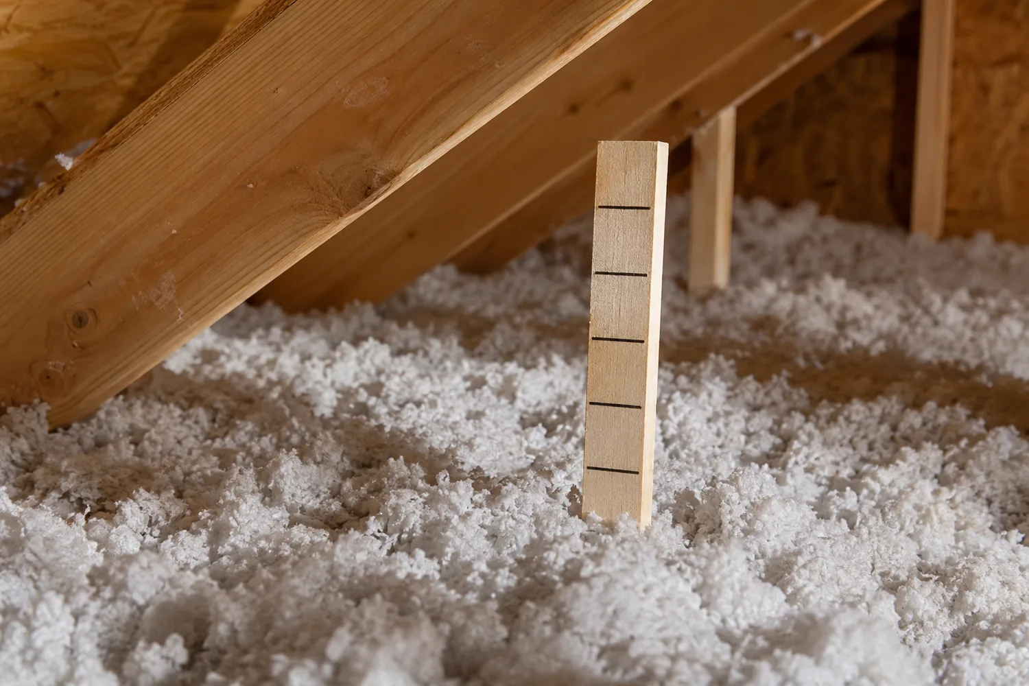 A wooden depth marker standing in loose-fill insulation beneath roof framing.